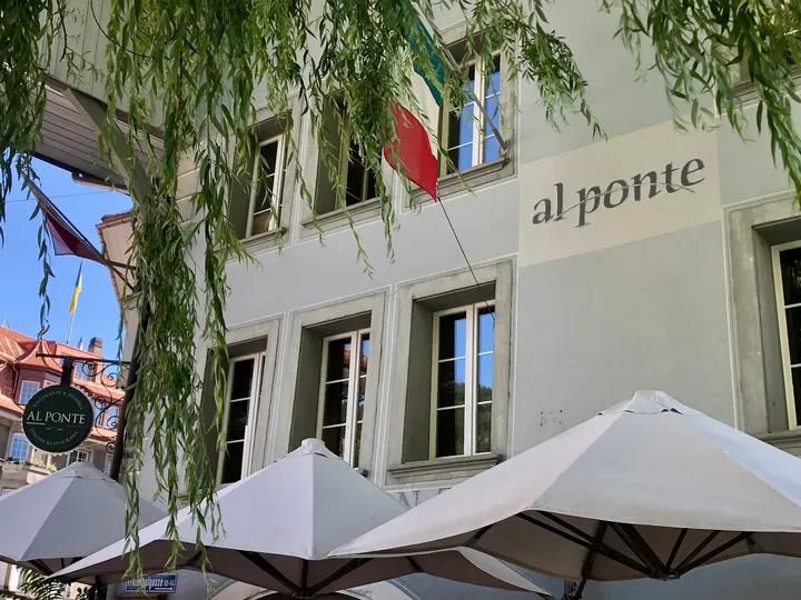 Die Fassade des Al Ponte mit Schriftzug, Sonnenschirmen und Trauerweide