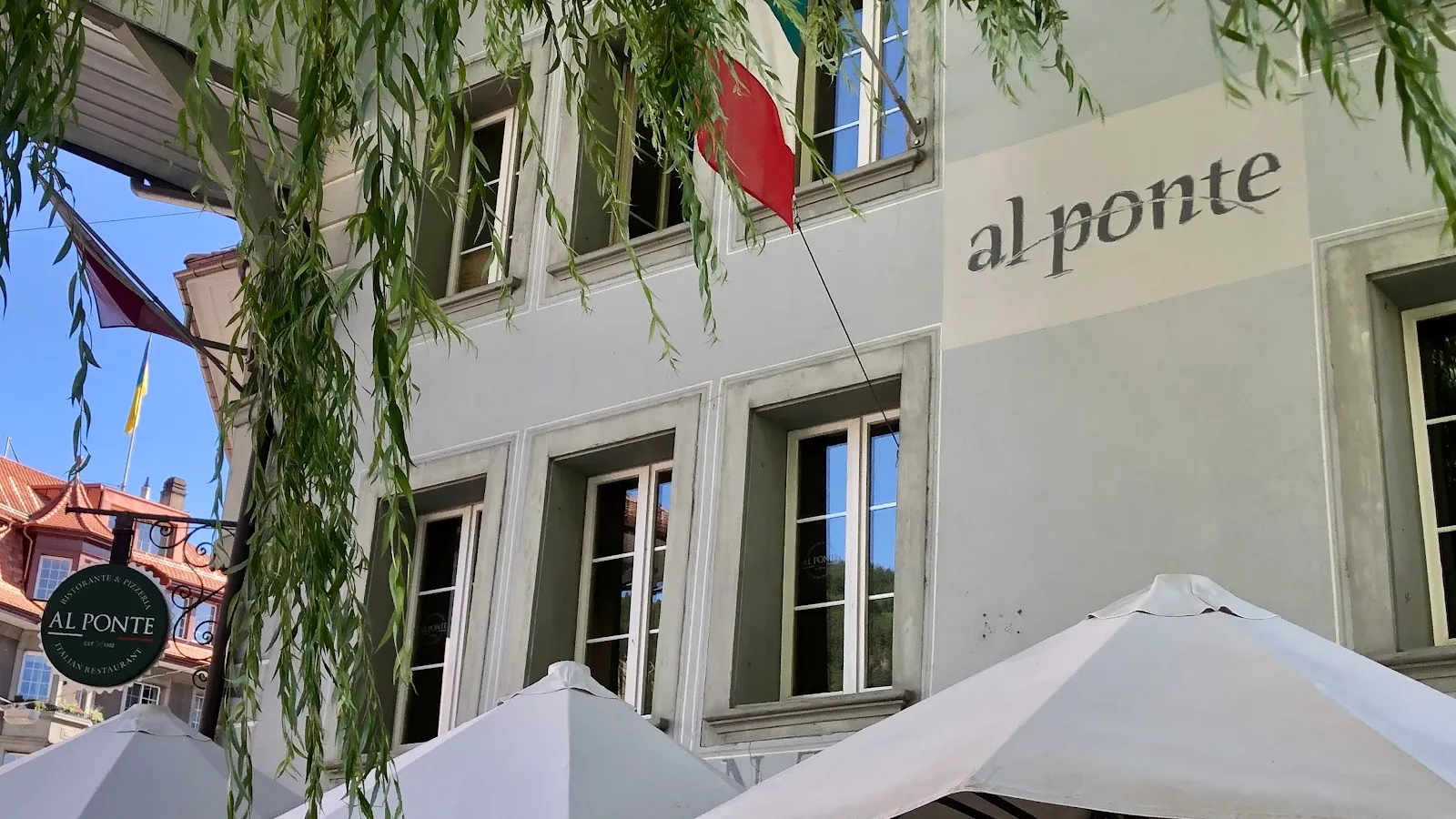 Die Fassade des Al Ponte mit Schriftzug, Sonnenschirmen und Trauerweide