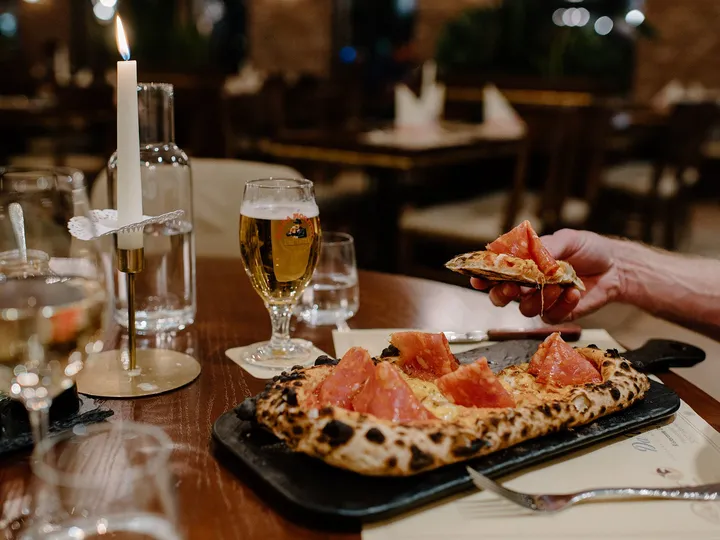Ein Stück Salami-Pizza vom Schieferbrett, dahinter ein Glas Bier