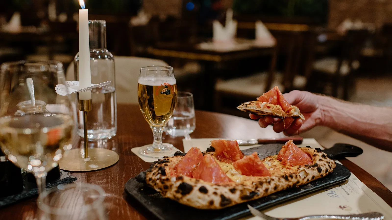 Ein Stück Salami-Pizza vom Schieferbrett, dahinter ein Glas Bier